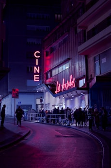 A nighttime scene featuring a cinema with neon lights. The marquee displays 'Los Ángeles,' and a crowd of people line up outside on the street. The area is illuminated by pink and purple lights, creating a vibrant atmosphere against the dark surroundings.