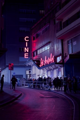 A nighttime scene featuring a cinema with neon lights. The marquee displays 'Los Ángeles,' and a crowd of people line up outside on the street. The area is illuminated by pink and purple lights, creating a vibrant atmosphere against the dark surroundings.