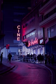 A nighttime scene featuring a cinema with neon lights. The marquee displays 'Los &Aacute;ngeles,' and a crowd of people line up outside on the street. The area is illuminated by pink and purple lights, creating a vibrant atmosphere against the dark surroundings.