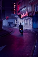 A motorcyclist rides along a curved city street at night, surrounded by neon lights. The neon signs display the words 'CINE' and 'Los Angeles', casting vivid reflections on the wet pavement. The urban setting is dimly lit, with cool and warm lighting creating a striking contrast.