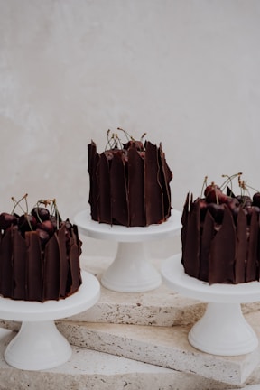 Three elegant chocolate cakes topped with cherries are displayed on white cake stands arranged on a textured stone surface, creating a sophisticated and minimalist presentation.