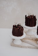 A set of elegant chocolate cakes adorned with cherries is displayed on white pedestals. The cakes are decorated with vertical pieces of chocolate and are placed on a stone slab with a minimalistic background.