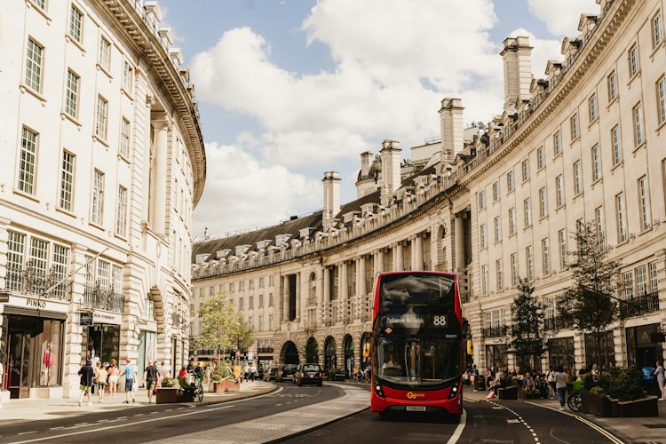 A red double-decker bus coming down the curve of Regent Street between cream stone terraces, with a black cab behind it and people on both pavements under a blue sky