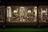 A beautifully decorated room with multiple Christmas trees adorned with lights, visible through large windows. Elegant wall sconces and mirrors add sophistication, while wreaths and garlands contribute to the festive atmosphere. The room features striped walls and neatly arranged chairs and tables.