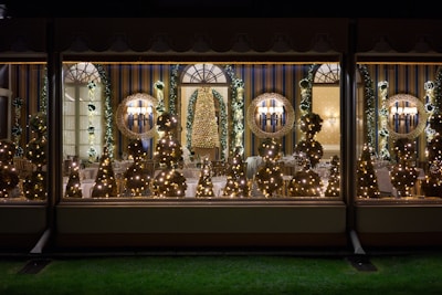 A beautifully decorated room with multiple Christmas trees adorned with lights, visible through large windows. Elegant wall sconces and mirrors add sophistication, while wreaths and garlands contribute to the festive atmosphere. The room features striped walls and neatly arranged chairs and tables.