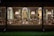 A beautifully decorated room with multiple Christmas trees adorned with lights, visible through large windows. Elegant wall sconces and mirrors add sophistication, while wreaths and garlands contribute to the festive atmosphere. The room features striped walls and neatly arranged chairs and tables.
