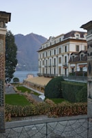 An elegant building with classic architecture is surrounded by neatly trimmed hedges and pathways illuminated with strings of lights. A large body of water and mountains provide a scenic backdrop. The outdoor space features a meticulously maintained garden with a white bench.