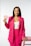 A person stands confidently against a plain backdrop wearing a bright pink blazer and pants with a white top. The person is holding two paper cards in one hand and resting the other hand on a pink stool. They have long dark hair and are smiling slightly.