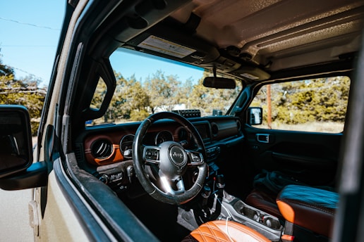 The interior of a Jeep is visible, featuring a leather-wrapped steering wheel and dashboard with detailed stitching on the seats. The door and part of the side mirror are in view, with a clear perspective of the vehicle's rugged, outdoor capability. Sunlight illuminates the interior, highlighting the textures and colors of the materials. Trees and greenery can be seen in the background through the windows.