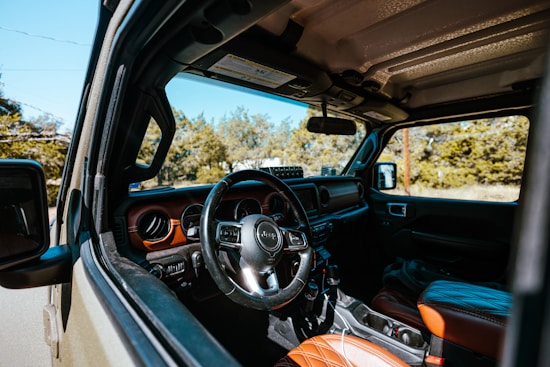 The interior of a Jeep is visible, featuring a leather-wrapped steering wheel and dashboard with detailed stitching on the seats. The door and part of the side mirror are in view, with a clear perspective of the vehicle's rugged, outdoor capability. Sunlight illuminates the interior, highlighting the textures and colors of the materials. Trees and greenery can be seen in the background through the windows.