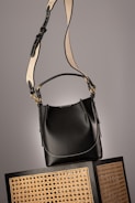 A black leather handbag with a beige and black adjustable strap hangs above two wicker chairs. The handbag features gold accents and white stitching, adding to its elegant design. The background is neutral, highlighting the bag and chairs.