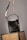 A black leather handbag with a beige and black adjustable strap hangs above two wicker chairs. The handbag features gold accents and white stitching, adding to its elegant design. The background is neutral, highlighting the bag and chairs.