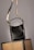 A black leather handbag with a beige and black adjustable strap hangs above two wicker chairs. The handbag features gold accents and white stitching, adding to its elegant design. The background is neutral, highlighting the bag and chairs.