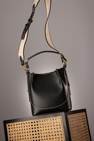 A black leather handbag with a beige and black adjustable strap hangs above two wicker chairs. The handbag features gold accents and white stitching, adding to its elegant design. The background is neutral, highlighting the bag and chairs.