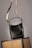 A black leather handbag with a beige and black adjustable strap hangs above two wicker chairs. The handbag features gold accents and white stitching, adding to its elegant design. The background is neutral, highlighting the bag and chairs.