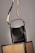 A black leather handbag with a beige and black adjustable strap hangs above two wicker chairs. The handbag features gold accents and white stitching, adding to its elegant design. The background is neutral, highlighting the bag and chairs.