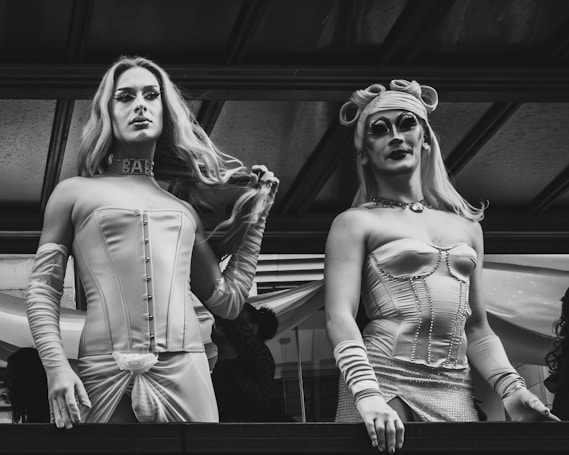 Two individuals dressed in elaborate, corseted outfits are standing side by side. Both have long hair and dramatic makeup, enhancing their expressive appearances. The outfits are stylish and feature detailed embellishments. The background suggests an indoor setting with a wooden ceiling structure.