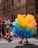 A person is dressed in a vibrant costume made entirely of multicolored balloons. The balloons form a large, colorful cluster around them. They stand amidst a crowd of people, who are casually dressed and appear to be enjoying an outdoor event. The setting is on a city street with a backdrop of a tall, old building.