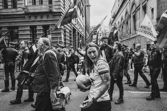 A black and white photo captures a group of people participating in a parade. Many individuals are dressed in leather attire, carrying flags that suggest a sense of identity and solidarity. Buildings frame the street, enhancing the urban setting.