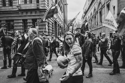 A black and white photo captures a group of people participating in a parade. Many individuals are dressed in leather attire, carrying flags that suggest a sense of identity and solidarity. Buildings frame the street, enhancing the urban setting.