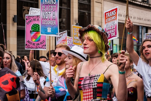 A lively group of people participating in a parade or march, holding colorful signs with messages supporting transgender rights. One sign reads 'Trans is a gift, not a disorder' while another states 'Trans lesbians are valid and really hot too.' The participants are dressed in vibrant clothing, with one person wearing a hat and sunglasses and another sporting neon green hair.