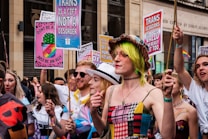 A lively group of people participating in a parade or march, holding colorful signs with messages supporting transgender rights. One sign reads 'Trans is a gift, not a disorder' while another states 'Trans lesbians are valid and really hot too.' The participants are dressed in vibrant clothing, with one person wearing a hat and sunglasses and another sporting neon green hair.