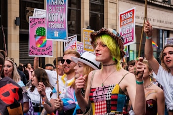 A lively group of people participating in a parade or march, holding colorful signs with messages supporting transgender rights. One sign reads 'Trans is a gift, not a disorder' while another states 'Trans lesbians are valid and really hot too.' The participants are dressed in vibrant clothing, with one person wearing a hat and sunglasses and another sporting neon green hair.