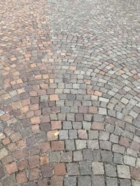 A detailed pattern of small cobblestones arranged in concentric arcs, creating a visually appealing surface with various shades of gray, brown, and reddish tones. A single dried leaf lies on the cobblestone, adding a touch of nature to the scene.