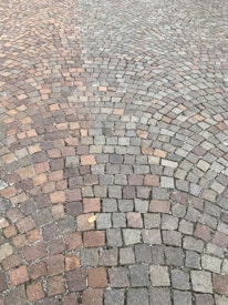 A detailed pattern of small cobblestones arranged in concentric arcs, creating a visually appealing surface with various shades of gray, brown, and reddish tones. A single dried leaf lies on the cobblestone, adding a touch of nature to the scene.
