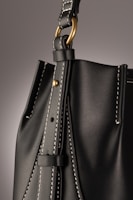 A close-up of a leather bag highlighting its textured dark material with white stitching. The bag features a gold-colored ring attaching the strap with visible threading details, giving an impression of finely crafted, high-quality design.