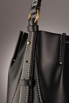 A close-up of a leather bag highlighting its textured dark material with white stitching. The bag features a gold-colored ring attaching the strap with visible threading details, giving an impression of finely crafted, high-quality design.