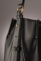 A close-up of a leather bag highlighting its textured dark material with white stitching. The bag features a gold-colored ring attaching the strap with visible threading details, giving an impression of finely crafted, high-quality design.