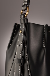A close-up of a leather bag highlighting its textured dark material with white stitching. The bag features a gold-colored ring attaching the strap with visible threading details, giving an impression of finely crafted, high-quality design.