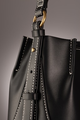 A close-up of a leather bag highlighting its textured dark material with white stitching. The bag features a gold-colored ring attaching the strap with visible threading details, giving an impression of finely crafted, high-quality design.