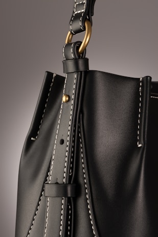 A close-up of a leather bag highlighting its textured dark material with white stitching. The bag features a gold-colored ring attaching the strap with visible threading details, giving an impression of finely crafted, high-quality design.