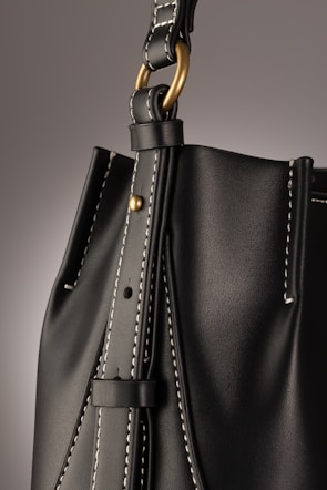 A close-up of a leather bag highlighting its textured dark material with white stitching. The bag features a gold-colored ring attaching the strap with visible threading details, giving an impression of finely crafted, high-quality design.
