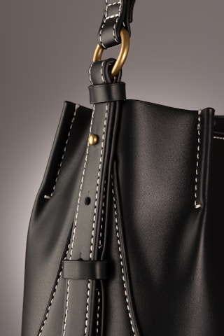 A close-up of a leather bag highlighting its textured dark material with white stitching. The bag features a gold-colored ring attaching the strap with visible threading details, giving an impression of finely crafted, high-quality design.