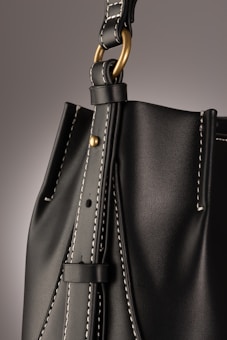 A close-up of a leather bag highlighting its textured dark material with white stitching. The bag features a gold-colored ring attaching the strap with visible threading details, giving an impression of finely crafted, high-quality design.