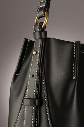 A close-up of a leather bag highlighting its textured dark material with white stitching. The bag features a gold-colored ring attaching the strap with visible threading details, giving an impression of finely crafted, high-quality design.