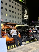 Two individuals are sitting on the open trunks of two parked cars, one orange and one black, in front of a building with the sign 'UMA Residences' illuminated. The setting appears to be at night, with the building's windows showing warm yellow lights. One person is holding an object while the other is using a phone, both casually dressed.
