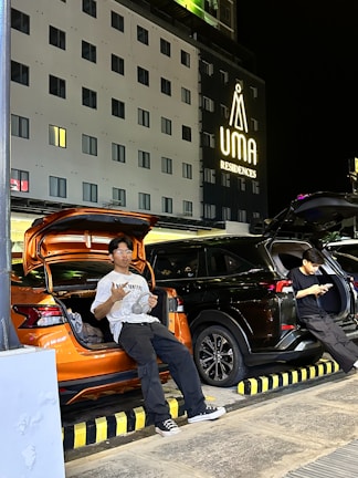 Two individuals are sitting on the open trunks of two parked cars, one orange and one black, in front of a building with the sign 'UMA Residences' illuminated. The setting appears to be at night, with the building's windows showing warm yellow lights. One person is holding an object while the other is using a phone, both casually dressed.