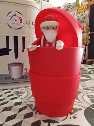 A toy figure dressed as Santa Claus is placed inside a red cup with a coffee machine in the background. The figure has a white beard, red hat, and is surrounded by a festive setting with decorative elements, including a patterned table surface.