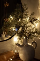 A ceramic jug with a spout and handle is placed on a wooden surface. Sprigs of greenery are arranged in the jug, illuminated by a warm string of fairy lights. In the background, a round mirror partially reflects the scene. Decorative metal bells hang from a string near the greenery.