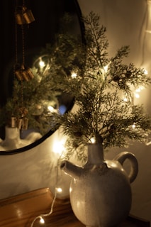 A ceramic jug with a spout and handle is placed on a wooden surface. Sprigs of greenery are arranged in the jug, illuminated by a warm string of fairy lights. In the background, a round mirror partially reflects the scene. Decorative metal bells hang from a string near the greenery.