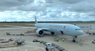 An aircraft with Air Canada branding is stationed at an airport gate with several workers surrounding it. The sky is overcast, and airport vehicles and equipment are present on the tarmac.