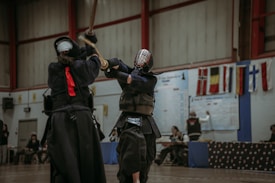 Two individuals are engaged in a kendo match, wearing traditional protective gear and wielding bamboo swords. The setting appears to be an indoor sports hall with various national flags on display in the background. Spectators and officials can be seen seated or standing around the edges.