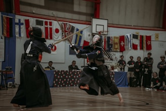 Two individuals are engaged in a kendo match, dressed in traditional kendo armor and wielding bamboo swords. They are in mid-action on a gymnasium floor, surrounded by spectators. Various international flags are displayed on the wall.