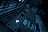 A close-up view of audio equipment with various knobs, buttons, and dials. The equipment is illuminated with a dim blue light, creating a moody atmosphere. Cables are plugged into the panel, indicating a setup in use or ready to be used.