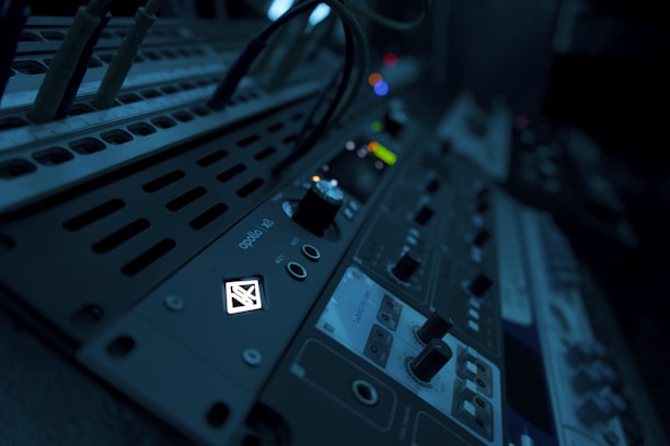 A close-up view of audio equipment with various knobs, buttons, and dials. The equipment is illuminated with a dim blue light, creating a moody atmosphere. Cables are plugged into the panel, indicating a setup in use or ready to be used.