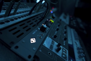 A close-up view of audio equipment with various knobs, buttons, and dials. The equipment is illuminated with a dim blue light, creating a moody atmosphere. Cables are plugged into the panel, indicating a setup in use or ready to be used.